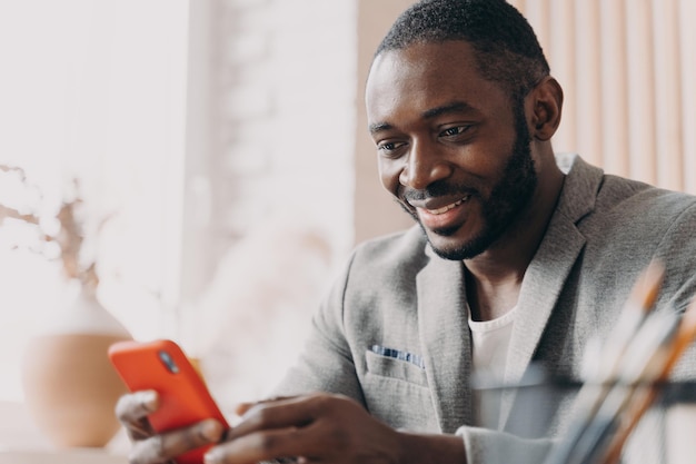 Uomo d'affari afroamericano sorridente che usa la chat mobile online mentre è seduto alla scrivania dell'ufficio
