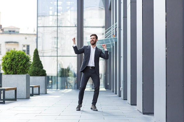 Uomo d'affari allegro felice che balla a piedi sullo sfondo della strada della città un moderno edificio per uffici fuori, all'aperto Divertente uomo d'affari di successo in tuta celebtes vittoria