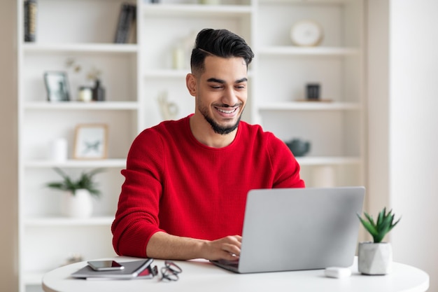 Uomo d'affari arabo millenario allegro e attraente con la barba in vestiti rossi che digita sul computer portatile in casa