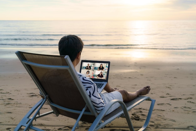 Uomo d'affari asiatico che ha una videoconferenza remota con il suo team di lavoro in spiaggia durante le vacanze in vacanza.
