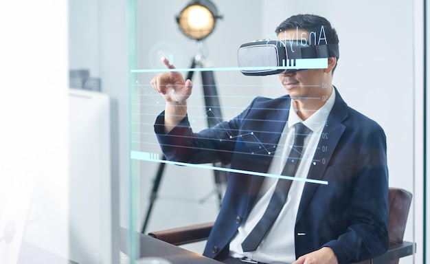Uomo d'affari asiatico che indossa occhiali di realtà virtuale e indica il grafico di crescita aziendale in un ufficio.