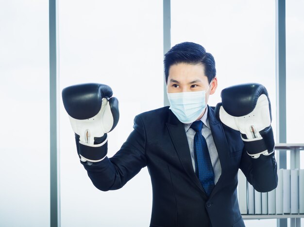 Uomo d'affari asiatico che indossa tuta e maschera protettiva e guantoni da boxe da portare mano in ufficio