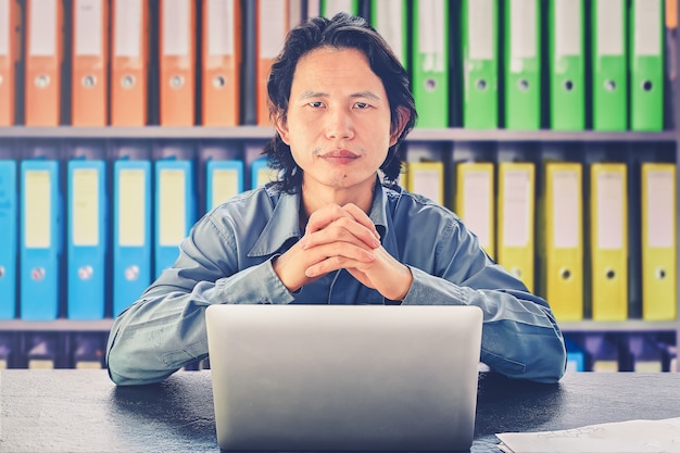 Uomo d'affari asiatico che per mezzo del computer portatile che si siede sullo sguardo dello scrittorio alla macchina fotografica