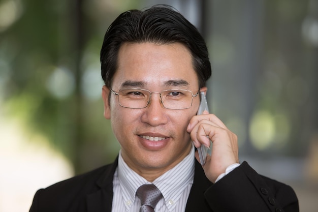 Uomo d'affari asiatico con gli occhiali in tuta parlando sul cellulare sorridente