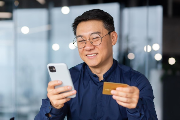 Uomo d'affari asiatico di successo e sorridente in ufficio che effettua bonifico bancario e acquisto online in