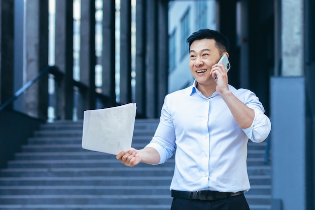 Uomo d'affari asiatico felice e di successo che parla al telefono tenendo i documenti fuori dall'ufficio