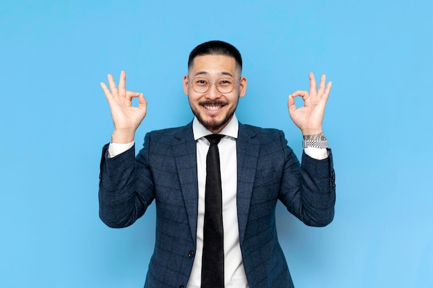uomo d'affari asiatico in giacca e occhiali mostra il gesto di ok su sfondo blu isolato