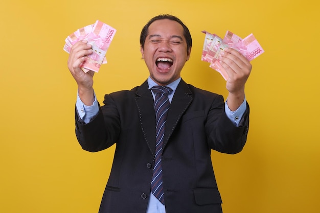 Uomo d'affari asiatico in tuta in piedi su sfondo giallo che celebra il successo e tiene denaro