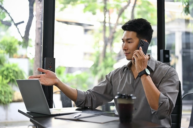 Uomo d'affari asiatico intelligente che guarda lo schermo del suo laptop e si occupa di affari al telefono