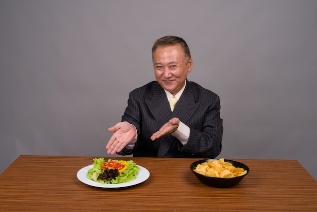 Uomo d'affari asiatico maturo che si siede con la tavola di legno contro gray