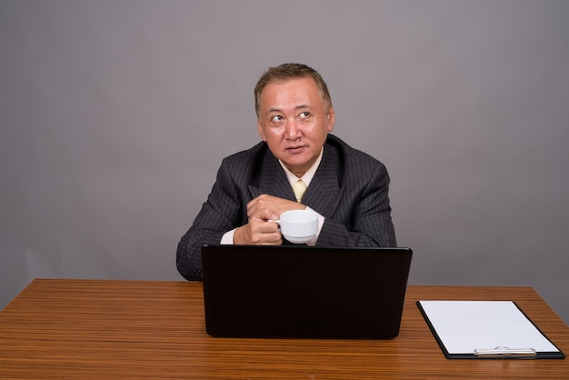 Uomo d'affari asiatico maturo che si siede con la tavola di legno contro il grigio