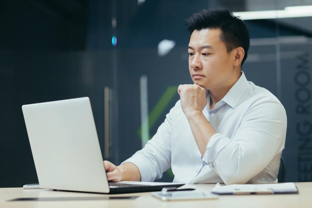 Uomo d'affari asiatico pensieroso e serio che lavora in ufficio pensando al proiettore che lavora con il laptop