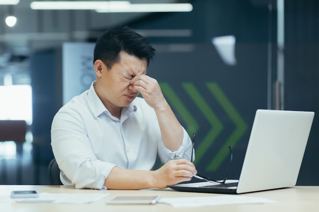 Uomo d'affari asiatico stanco che lavora fino a tardi negli occhi irritati dell'ufficio uomo con il computer portatile