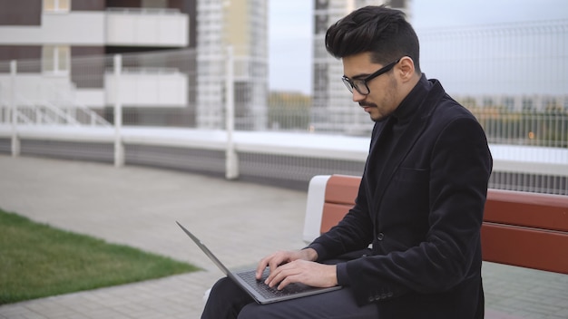 Uomo d'affari attraente in abito nero con laptop seduto all'aperto, copyspace