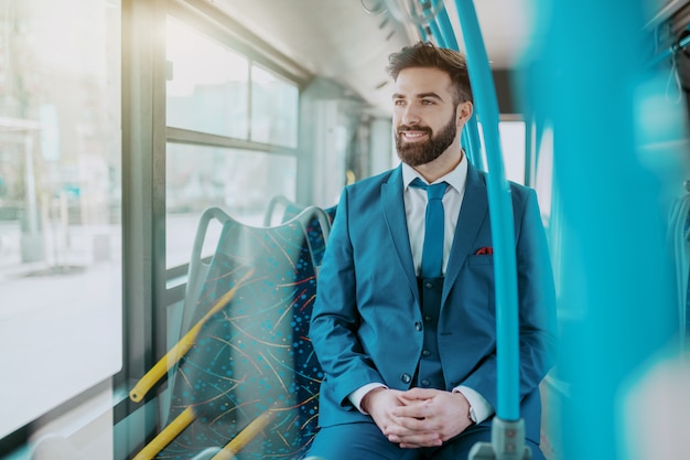 Uomo d'affari attraente sorridente dei giovani in vestito blu che si siede in bus pubblico e che guarda la finestra della depressione.