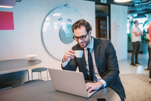 Uomo d'affari barbuto bello caucasico felice in vestito e con gli occhiali seduto al bancone della cucina in compagnia, bevendo caffè e usando il computer portatile.