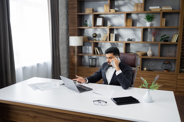 Uomo d'affari barbuto che lavora al computer portatile mentre fa una telefonata in un ufficio moderno