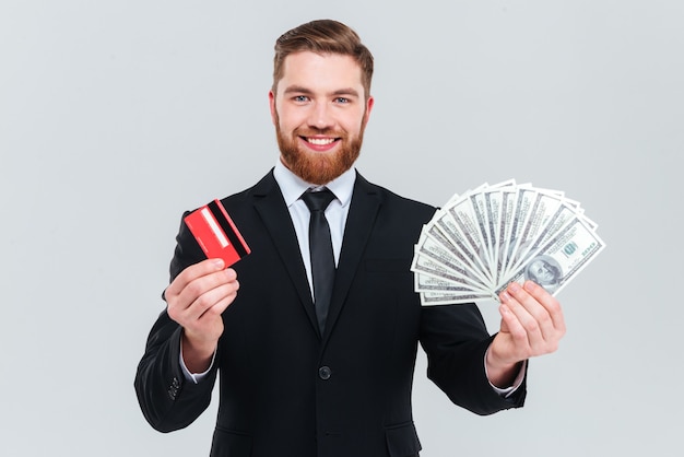 Uomo d'affari barbuto felice in abito nero con carta di credito e denaro in mano. Sfondo grigio isolato