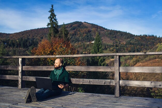 Uomo d'affari barbuto seduto sulla terrazza in montagna con il computer portatile e parlando al telefono