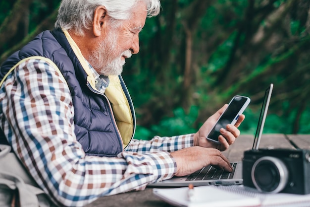 Uomo d'affari barbuto sorridente che lavora al computer portatile guardando il telefono seduto all'aperto nel parco