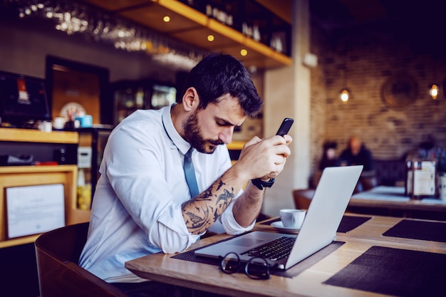 Uomo d'affari barbuto tatuato caucasico serio in camicia e cravatta che si siede nella caffetteria, che tiene telefono astuto e pensiero. Sul tavolo davanti a lui c'è il laptop.