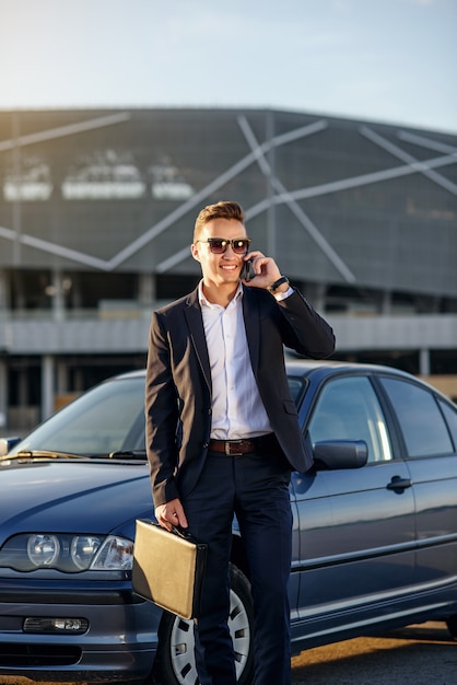 Uomo d'affari bello attraente con il diplomatico che parla sullo smartphone vicino all'automobile