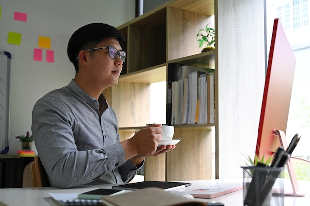 Uomo d'affari bello che beve tazza di caffè e lavora con il computer