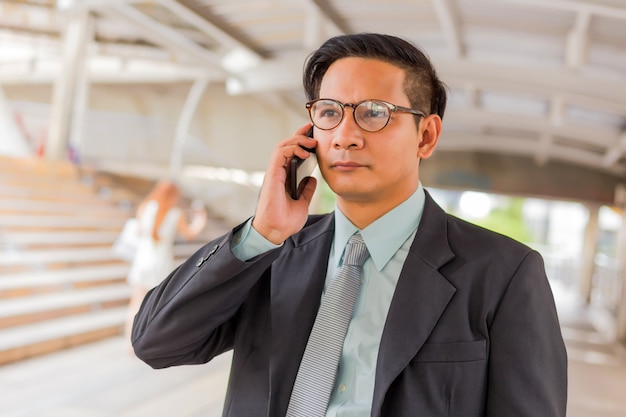 Uomo d'affari bello del giovane Asia con il suo smartphone che sta sul passaggio pedonale della città moderna.