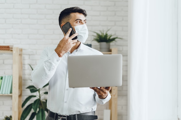 Uomo d'affari bello dell'uomo nella mascherina medica usando il suo computer portatile mentre parla sul suo cellulare all'interno