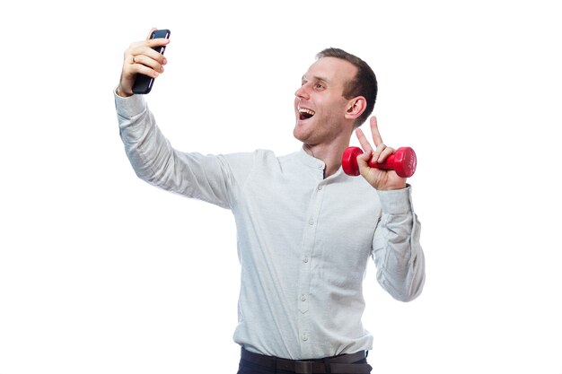 Uomo d'affari caucasico, un insegnante fa selfie telefono e sport. Ha in mano un manubrio rosso. Indossa una maglietta. Ritratto emotivo. Isolato su sfondo bianco