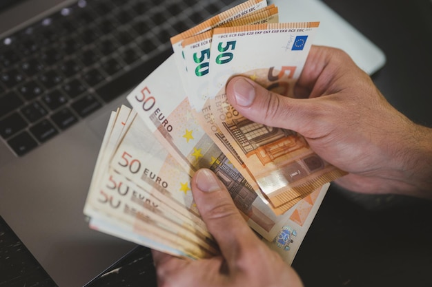 Uomo d'affari che conta soldi. le mani maschili ricche tengono e contano banconote in contanti di banconote da 50 euro o banconote in valuta davanti a un laptop