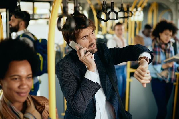 Uomo d'affari che controlla il tempo sull'orologio da polso mentre parla al telefono in un autobus