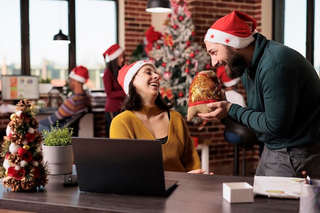 Uomo d'affari che dà scatola regalo alla donna in ufficio, celebrando le vacanze della vigilia di Natale con i regali. I colleghi si sentono felici per le festività natalizie con ornamenti e decorazioni natalizie.