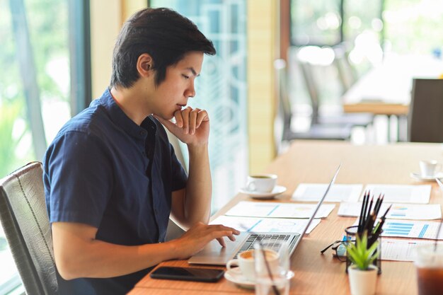 Uomo d'affari che guarda un laptop con relazione finanziaria, forniture per ufficio e tazza di caffè nello spazio di lavoro