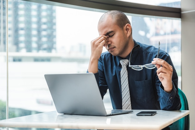 Uomo d'affari che ha emicrania e che lavora al computer portatile in ufficio con la costruzione della città