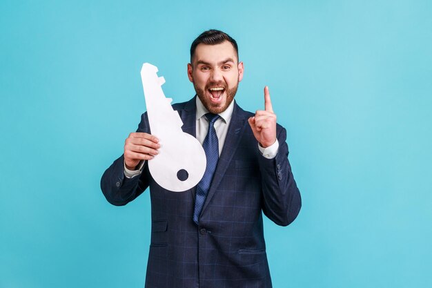 Uomo d'affari che indossa un abito in stile ufficiale che punta il dito verso l'alto e tiene in mano una grande chiave di carta soddisfatto dell'affitto o dell'acquisto di alloggi Studio indoor girato isolato su sfondo blu