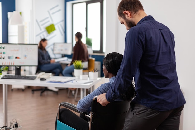 Uomo d'affari che porta una donna d'affari afroamericana handicappata paralizzata al lavoro nell'ufficio aziendale di avvio attività. Lavoro di squadra che lavora alla strategia di gestione che analizza le statistiche dell'azienda