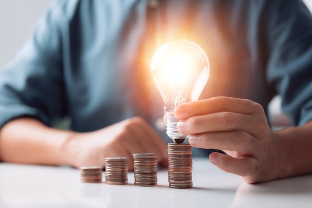Uomo d'affari che tiene e mette una lampadina sulla pila di monete per risparmiare energia e denaro concept