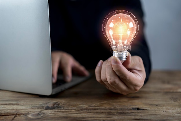 Uomo d'affari che tiene lampadina incandescente sulla tavola di legno con il computer portatile come soluzione aziendale