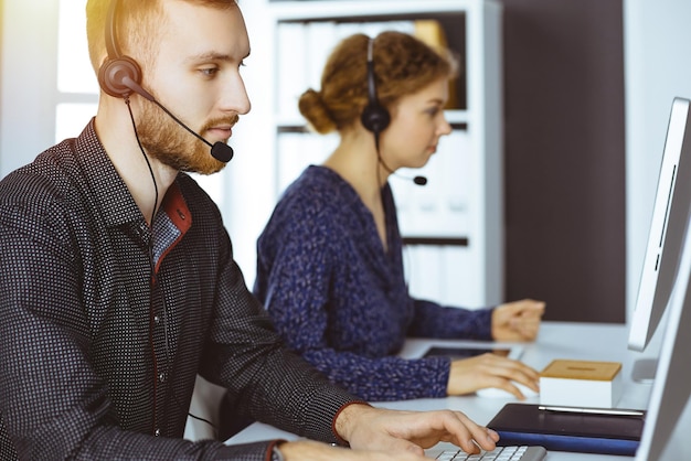 Uomo d'affari con la barba rossa che parla con l'auricolare vicino alla sua collega mentre è seduto in un moderno ufficio soleggiato. Gruppo di persone diverse nel call center.