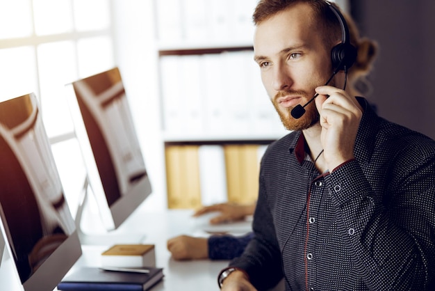 Uomo d'affari con la barba rossa che parla con l'auricolare vicino alla sua collega mentre è seduto in un moderno ufficio soleggiato. Gruppo di persone diverse nel call center.