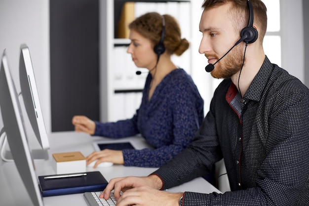 Uomo d'affari con la barba rossa che parla con l'auricolare vicino alla sua collega mentre è seduto in un ufficio moderno. Gruppo di persone diverse nel call center. Telemarketing e servizio clienti.