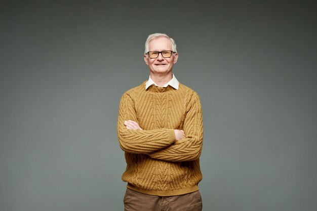 Uomo d'affari dai capelli grigi invecchiato contemporaneo in maglione marrone lavorato a maglia tenendo le braccia incrociate sul petto e guardando la fotocamera