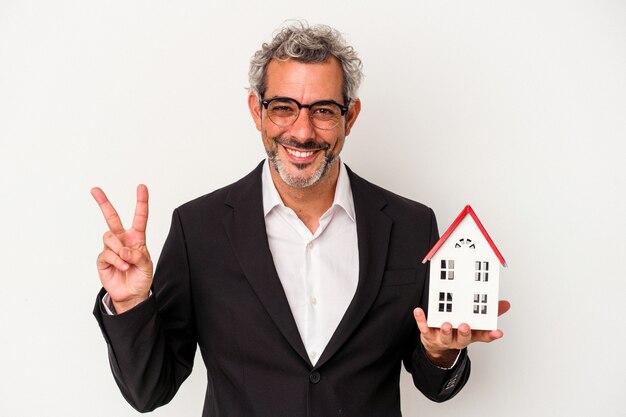 Uomo d'affari di mezza età che tiene fatture e modello di casa isolato su sfondo blu che mostra il numero due con le dita.