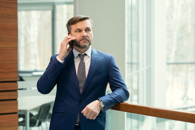 Uomo d'affari di mezza età serio e premuroso con stoppie appoggiato alla ringhiera nel corridoio dell'ufficio e parlando al telefono