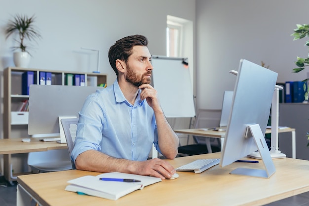 Uomo d'affari di pensiero che lavora in un ufficio moderno durante il giorno seduto a un tavolo che lavora con un computer pensando di risolvere un contratto commerciale