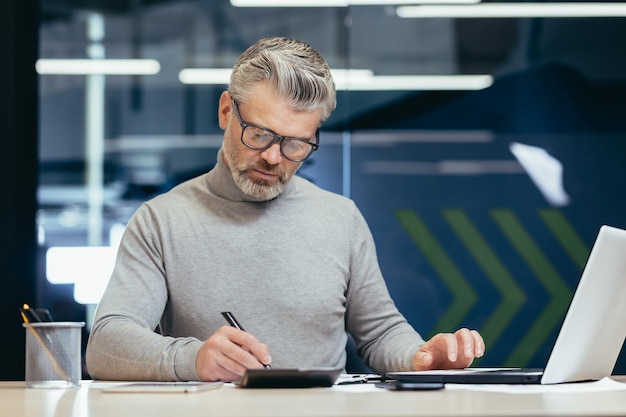 Uomo d'affari di pensiero serio all'interno dell'ufficio al lavoro con il computer portatile uomo dai capelli grigi senior che lavora