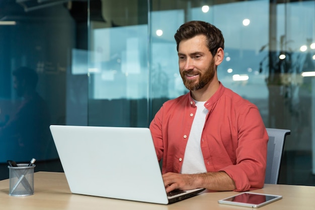 Uomo d'affari di successo in camicia rossa che lavora felicemente con il computer portatile all'interno dell'ufficio uomo maturo con la barba