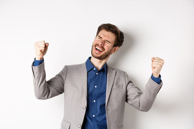 Uomo d'affari di successo in vestito che vince il premio, gridando sì con un sorriso sollevato e pompe a pugno, celebrando il successo e il successo, in piedi su sfondo bianco.