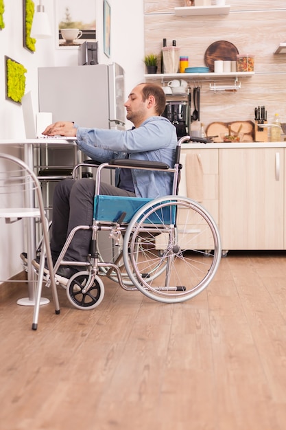 Uomo d'affari disabile in sedia a rotelle utilizzando il computer portatile in cucina. Disabile paralizzato uomo handicappato con disabilità motoria che si integra dopo un incidente.